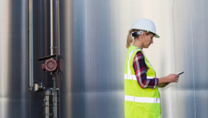 a female engineer in a high vis jacket and a hard hart in front of a large silver tank performing health and safety assessments